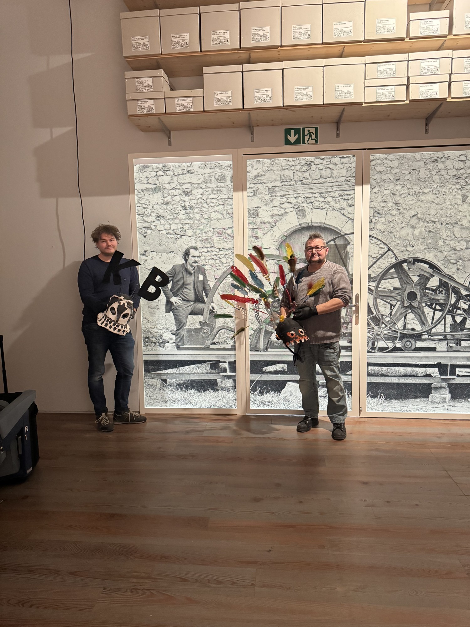 Jean-Marc Gaillard und Andy Hofmann posieren mit Fasnachtslarven vor Foto von Tinguely und Skulptur1@simonematthieu_paint