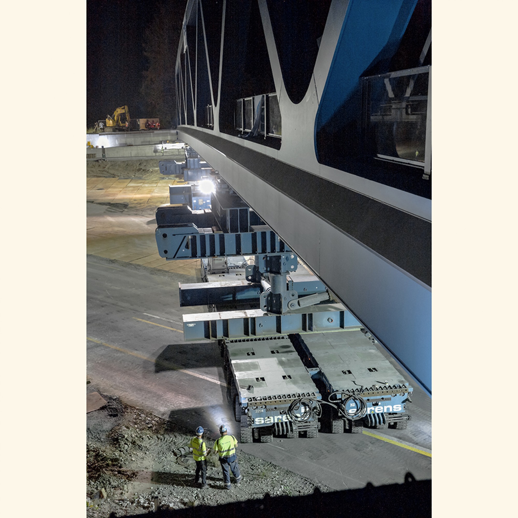 Das Einfahren der nördlichen Brücke mittels SPMT. Die Arbeiter mit den Warnwesten steuern mit Funkfernsteuerungen einmal die vordere und einmal die hintere Einheit von je 8 zusammen geschalteten Fahrzeugen.